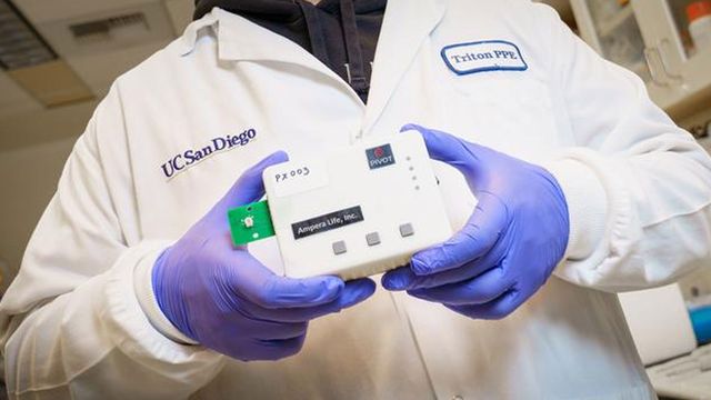 A scientist in gloves and a lab coat holds a small white device. 