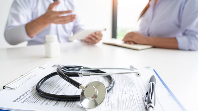 A form and stethoscope sit on a table, with two doctors in the background. 