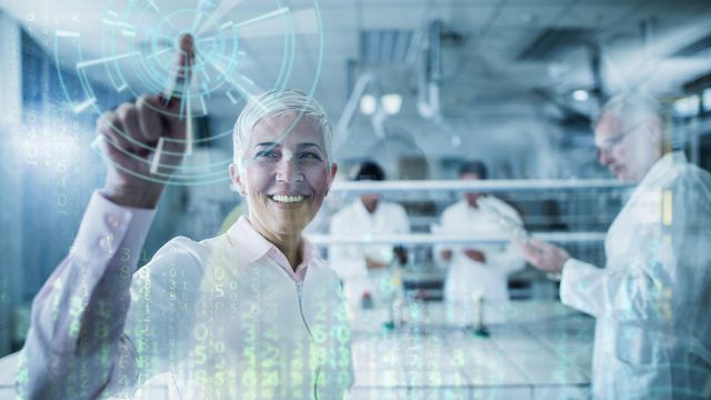 A smiling woman with short white hair, wearing a long-sleeved pink shirt stands in a lab, with her right arm raised and index finger touching a clear digital screen. In the background, three other out of focus people can be seen in the lab wearing white lab coats. 