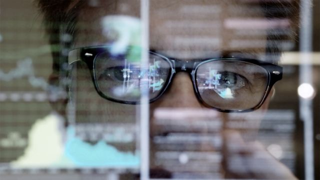 A man wearing glasses with black frames looks through a screen with a digital overlay. 