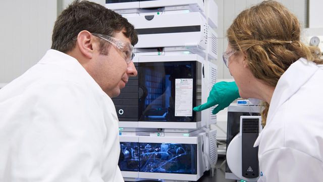 Two people in lab coats looking at an ACT label on a piece of analytical chemistry equipment. 