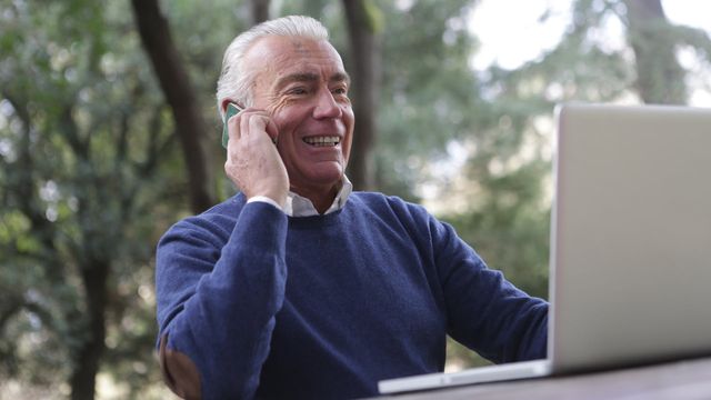 A man sits and talks on the phone while using a laptop. 